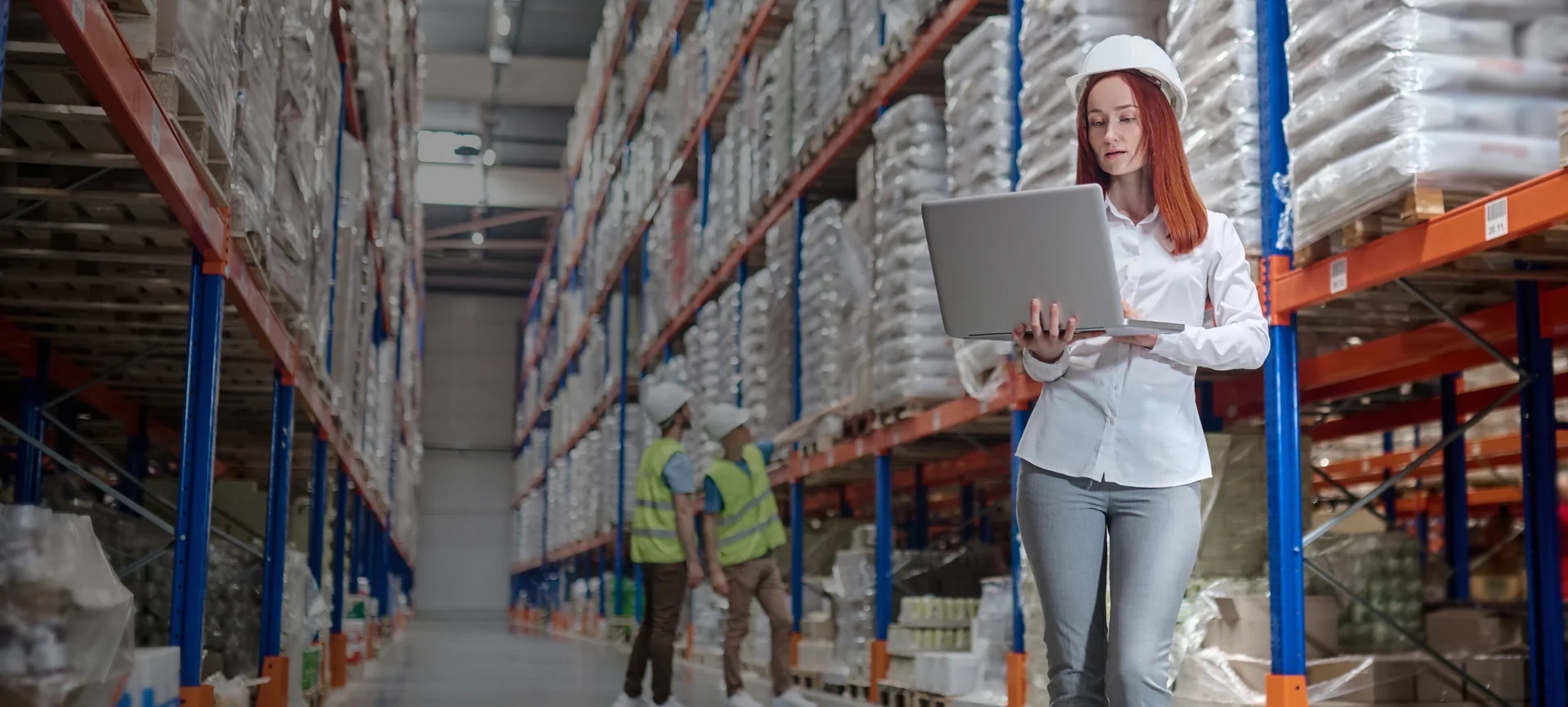 A woman in a white shirt and hat stands in a warehouse, working on a laptop, representing tech solutions for logistics.